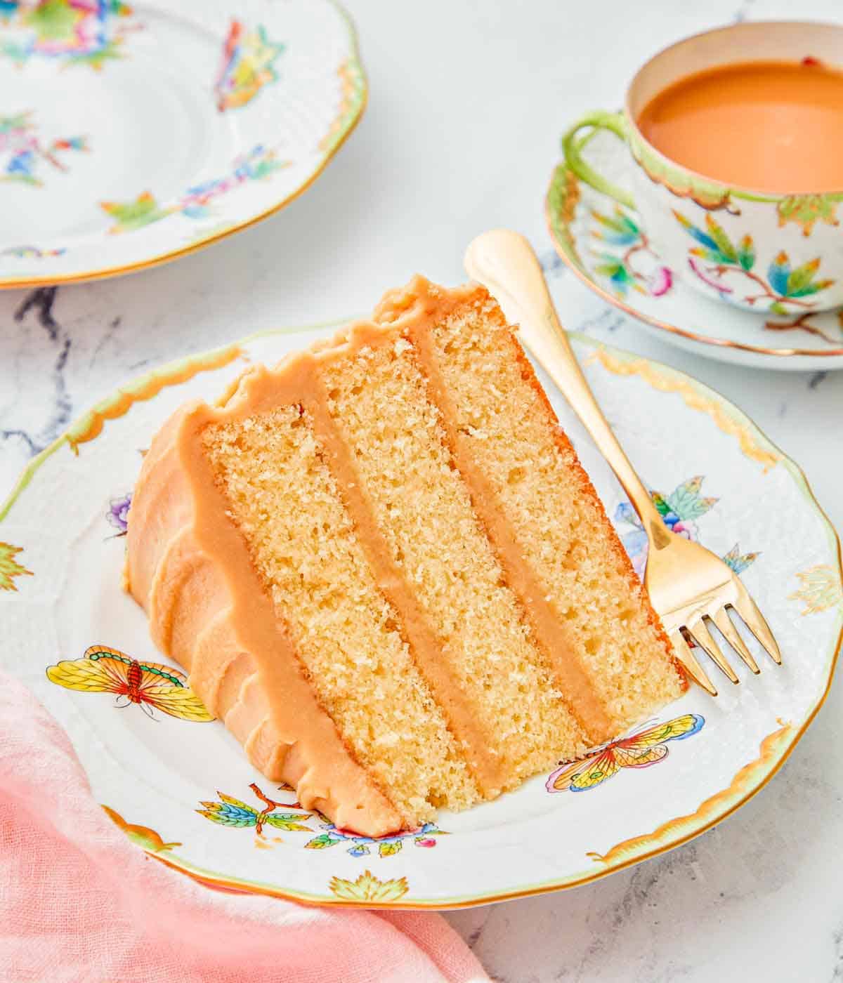 A slice of caramel cake on a plate with a fork, showing the three layers and frosting inbetween.