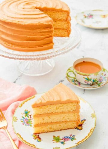 A slice of caramel cake on a plate with the rest of the cake on a stand in the background along with coffee.