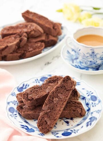 A plate with multiple chocolate biscottis with a cup of coffee and more biscotti in the background.