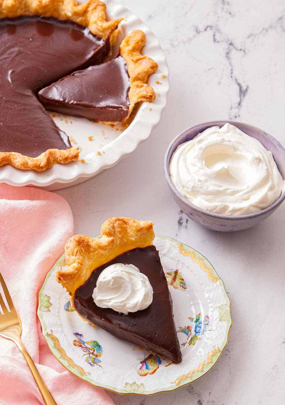A slice of chocolate pie with a bowl of whipped cream and the rest of the pie in the background.