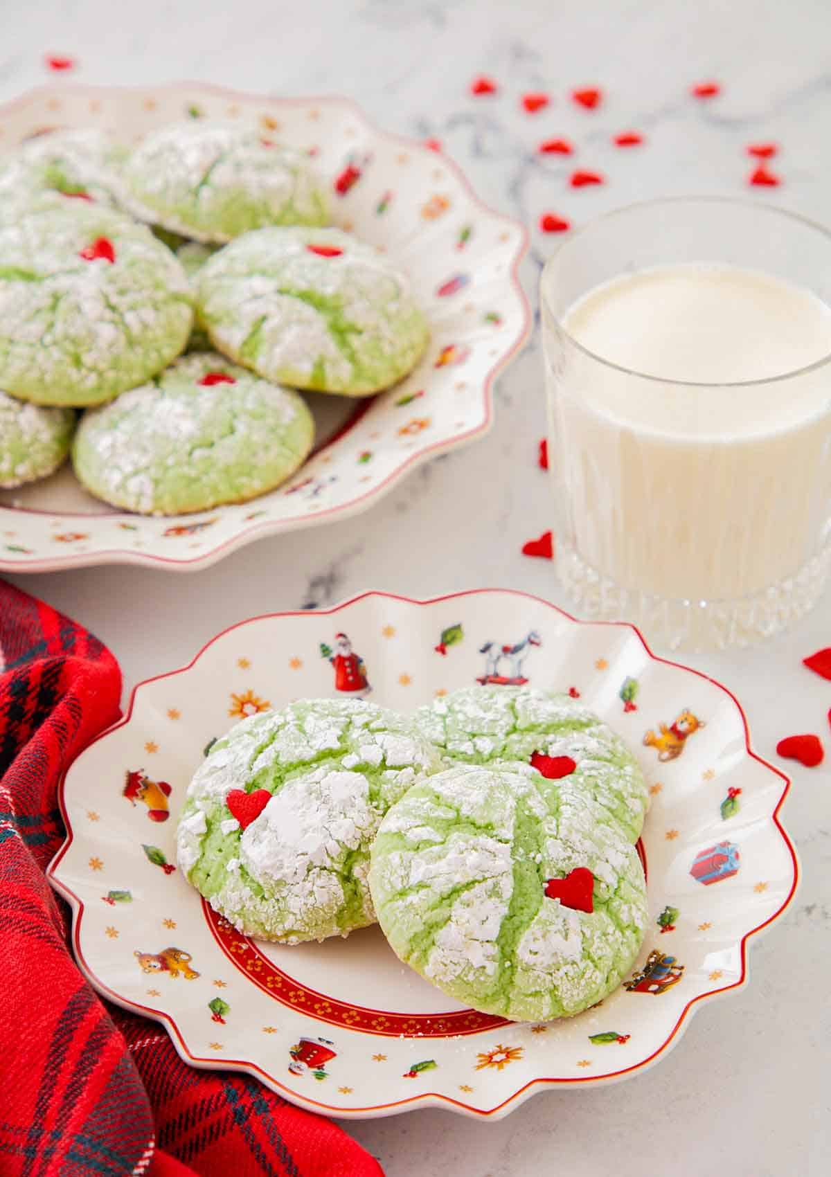 A plate with three Grinch cookies with a glass of milk and additional cookies in the background.