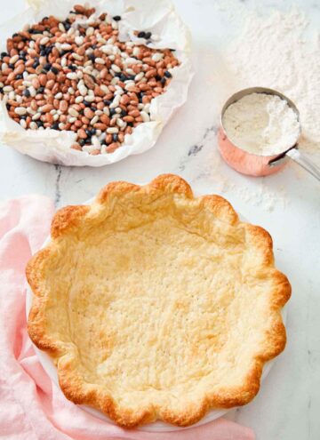 A baked pie crust with dried beans in parchment and a measuring cup of flour in the background.