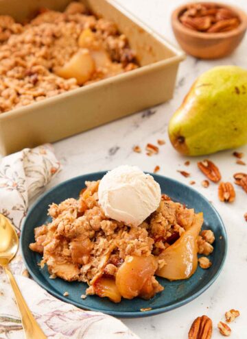 A plate of pear crisp with a scoop of vanilla ice cream on top with the rest of the baking dish in the back alongside a pear.