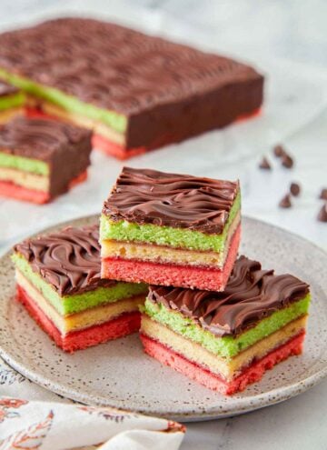 A plate with three pieces of rainbow cookies in a pyramid. The rest of the dessert in the background.