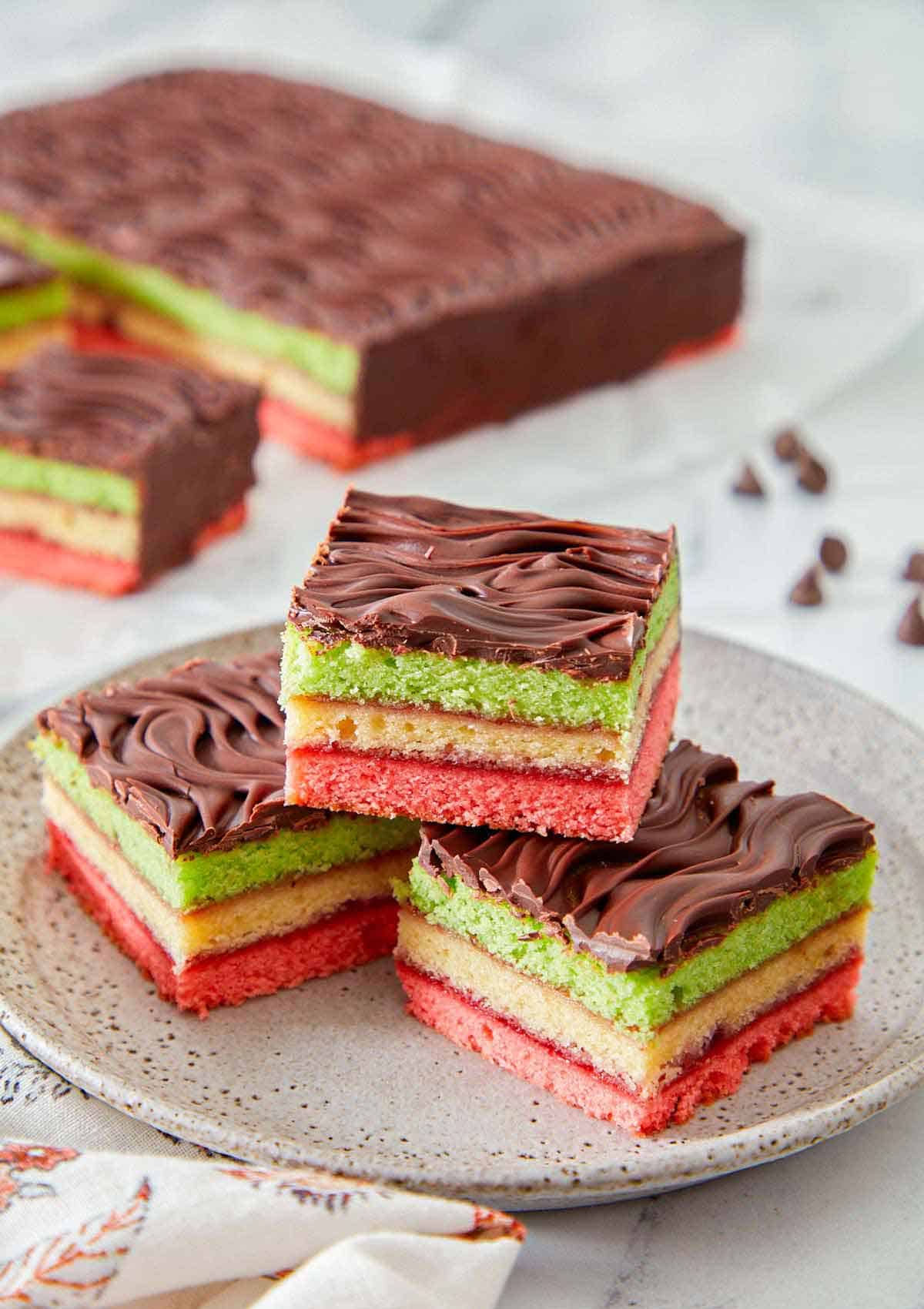 A plate with three pieces of rainbow cookies in a pyramid. The rest of the dessert in the background.