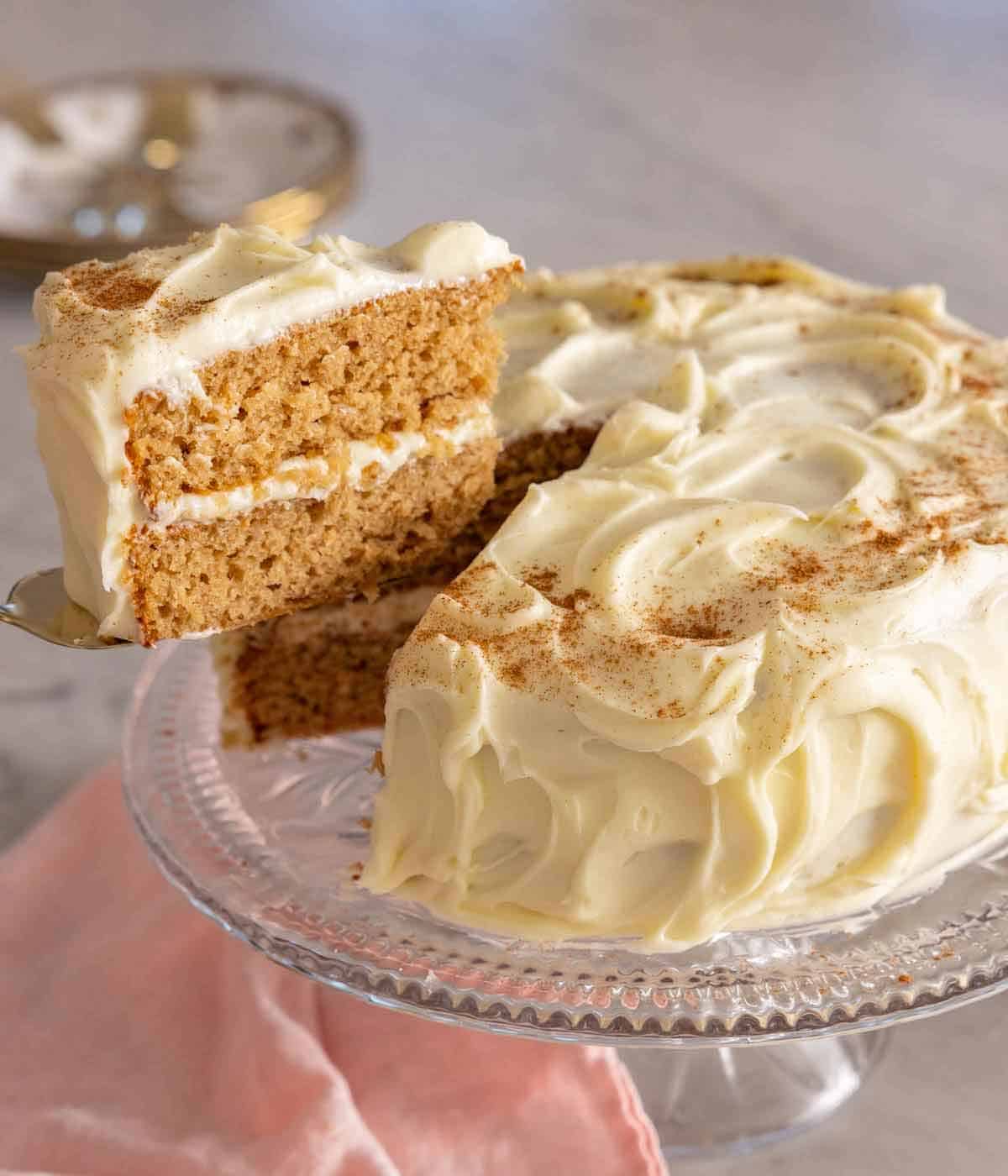 A slice of spice cake cut and lifted from the whole cake in a clear cake stand.