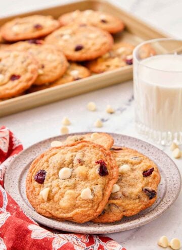 A plate with a couple of white chocolate cranberry cookies with a glass of milk and tray of cookies in the background.
