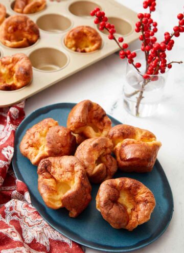 A blue platter with five Yorkshire puddings with a plant and more Yorkshire pudding in the background.