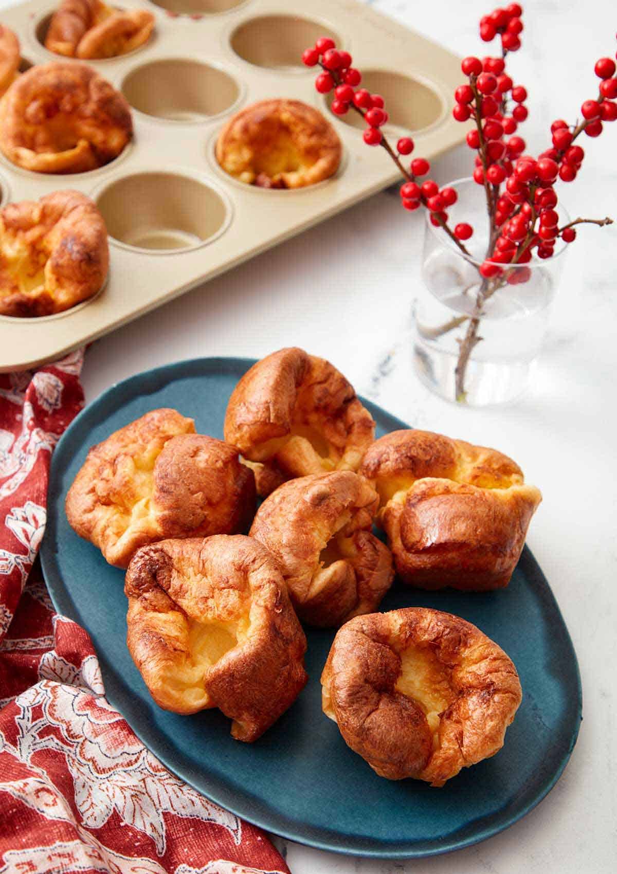 A blue platter with five Yorkshire puddings with a plant and more Yorkshire pudding in the background.