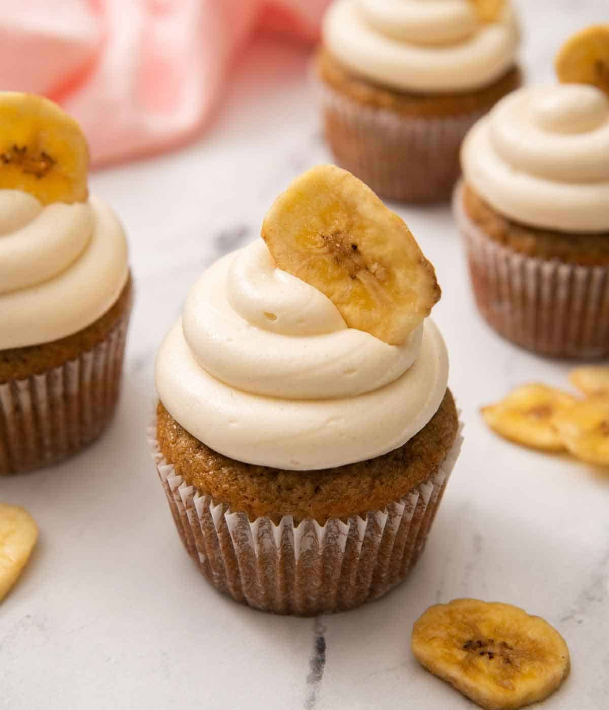 Multiple banana cupcakes with one in focus in the middle with a banana chip pressed into the frosting.