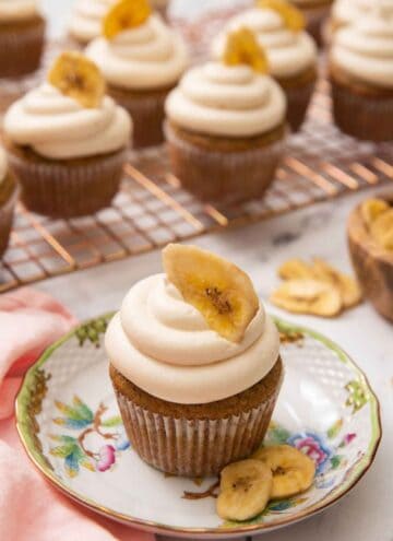 A small plate with a banana cupcake with a banana chip on top and more cupcakes in the back on a cooling rack.