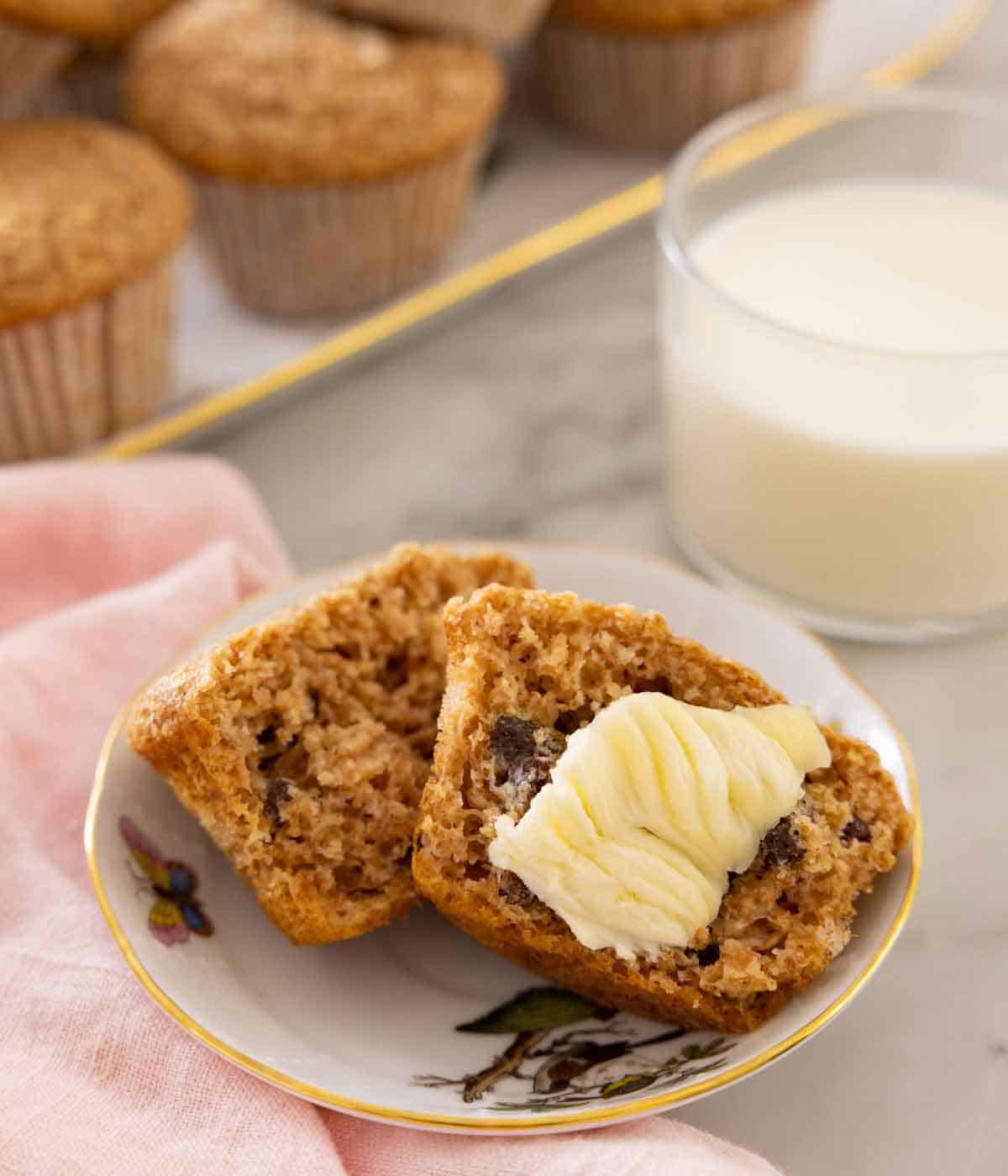 A plate with a bran muffin cut open in half with butter spread onto one half. A glass of milk in the back.