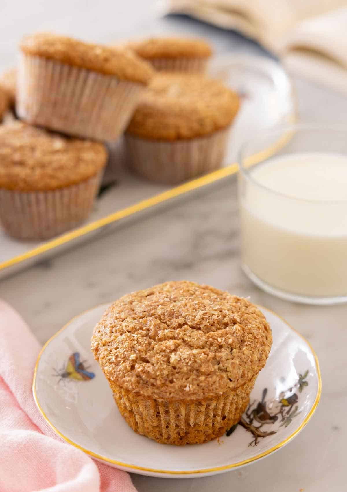 A small plate with a bran muffin with a glass of milk and another platter of muffins in the back.