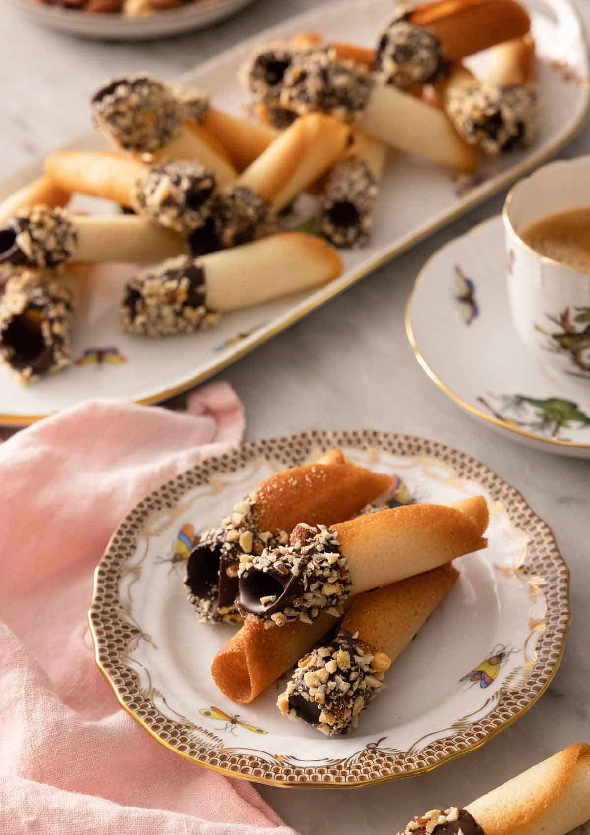 A plate with multiple chocolate dipped tuiles with a platter in the background with additional cookies.
