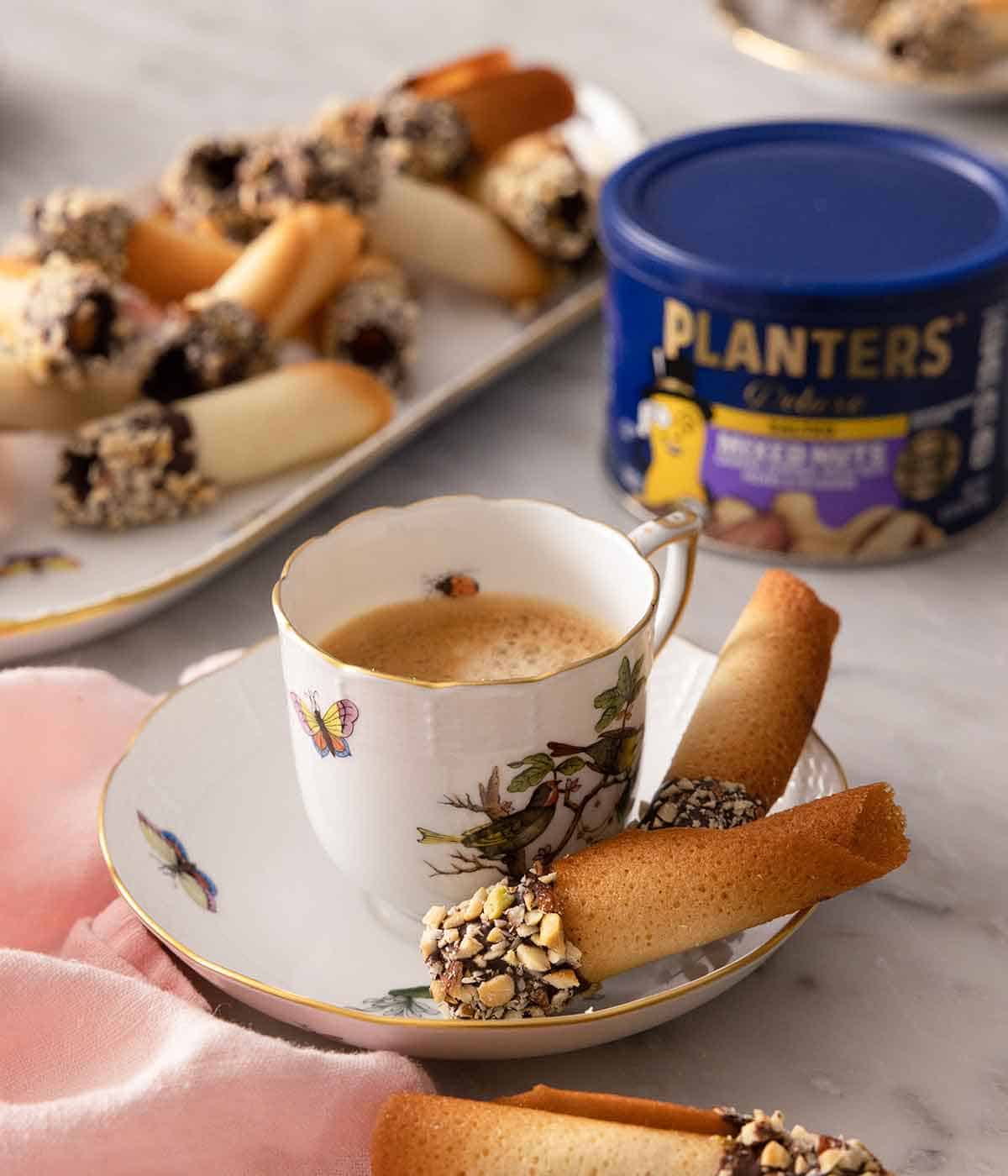 A mug of coffee with two chocolate dipped tuiles on the plate under the mug with more cookies in the back along with a container of nuts.