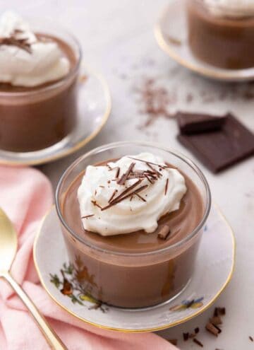 A glass of chocolate pudding with whipped cream and shaved chocolate on top with two more, out of focus, in the background.