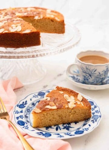 A plate with a slice of honey cake with the rest of the cake and a coffee in the background.