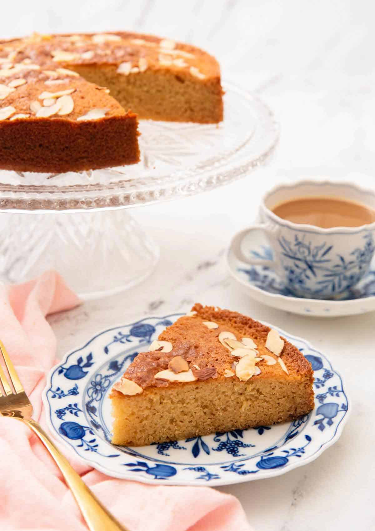 A plate with a slice of honey cake with the rest of the cake and a coffee in the background.