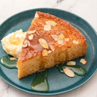 A plate with a slice of honey cake with honey drizzled on top and whipped cream beside it.