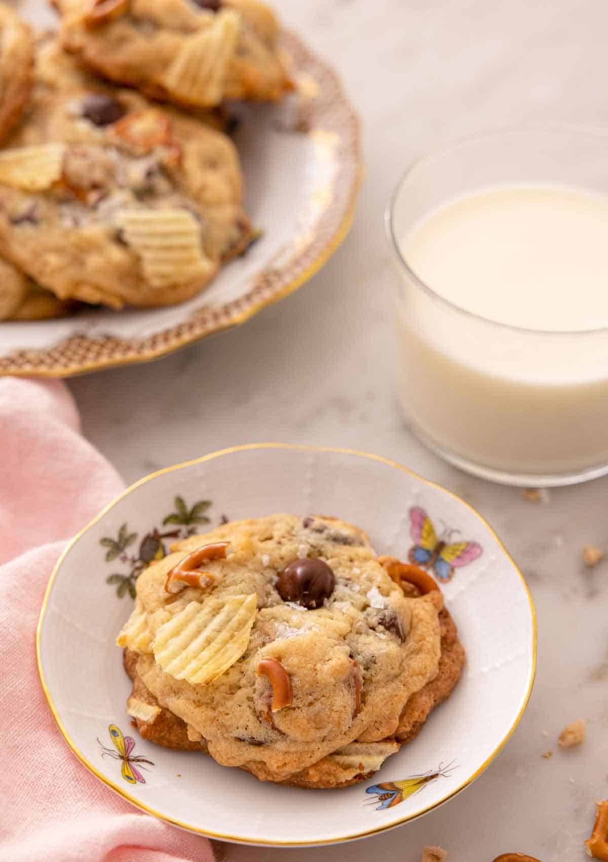 A small plate with a kitchen sink cookie with a glass on milk and a platter of more cookies behind it.