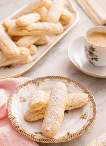 A plate with three ladyfingers with a cup of coffee and a tray of additional ladyfingers in the back.