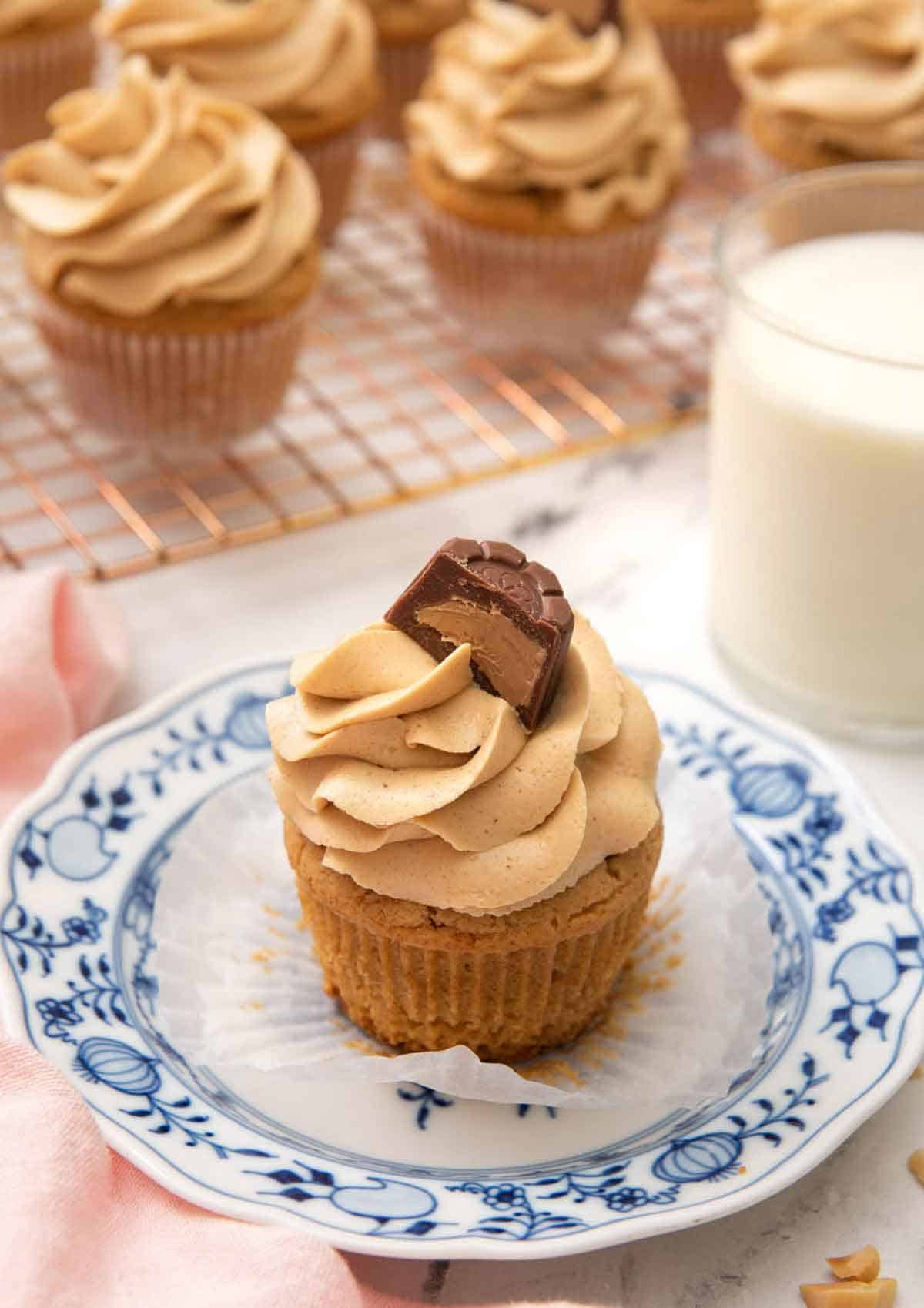 A small plate with a peanut butter cupcake with the liner pulled down and glass of milk and more cupcakes in the back.