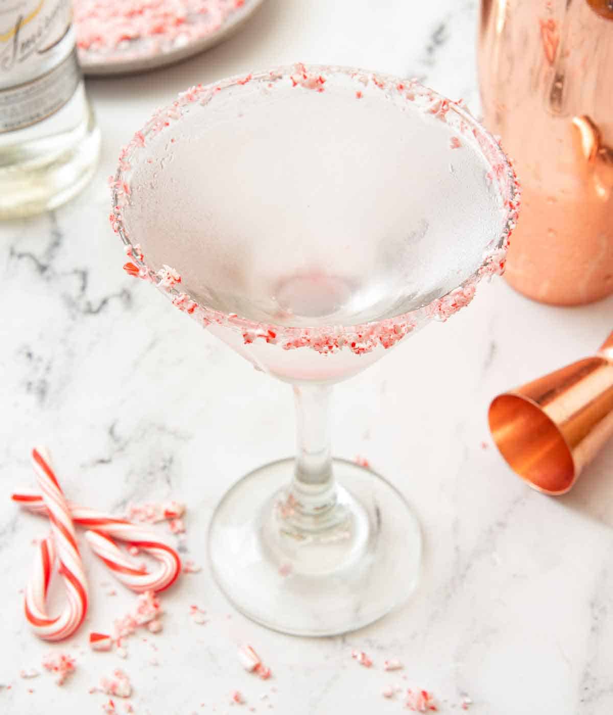 A peppermint martini with crushed and full peppermint candies scattered around with a jigger and shaker behind it.