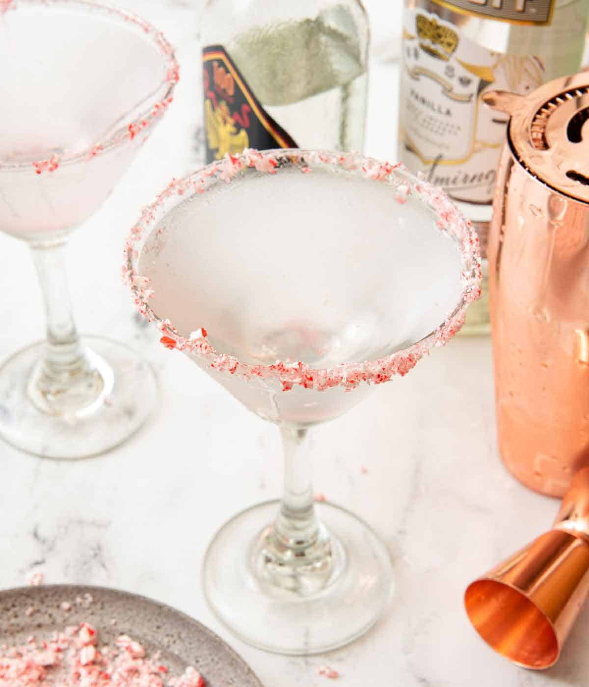 A glass of peppermint martini with a second glass, bottles of liqueur, and the shaker behind it.