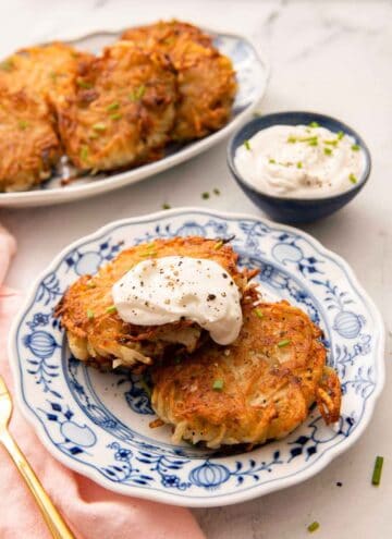 A plate with two potato pancakes with a dollop of sour cream on top. More pancakes and dip in the background.