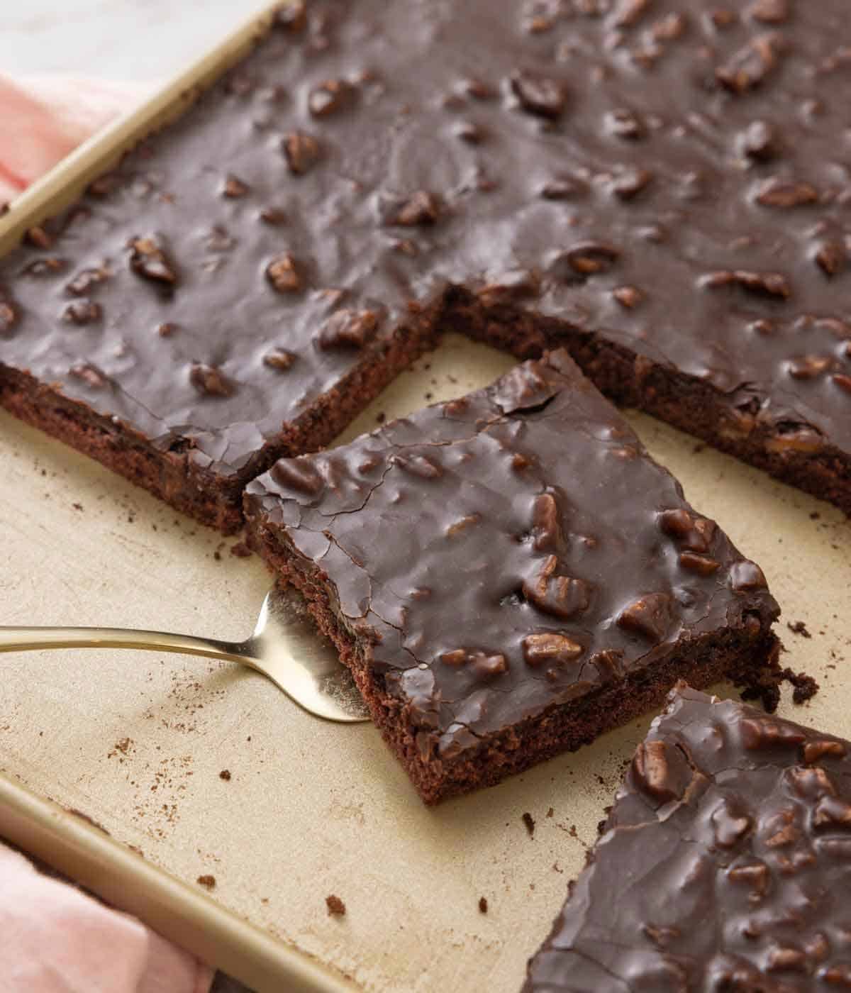 A sheet pan of Texas sheet cake with slices cut out and one with a spatula underneath it.