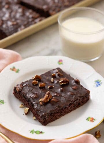A plate with a square slice of Texas sheet cake with a glass on milk in the back.