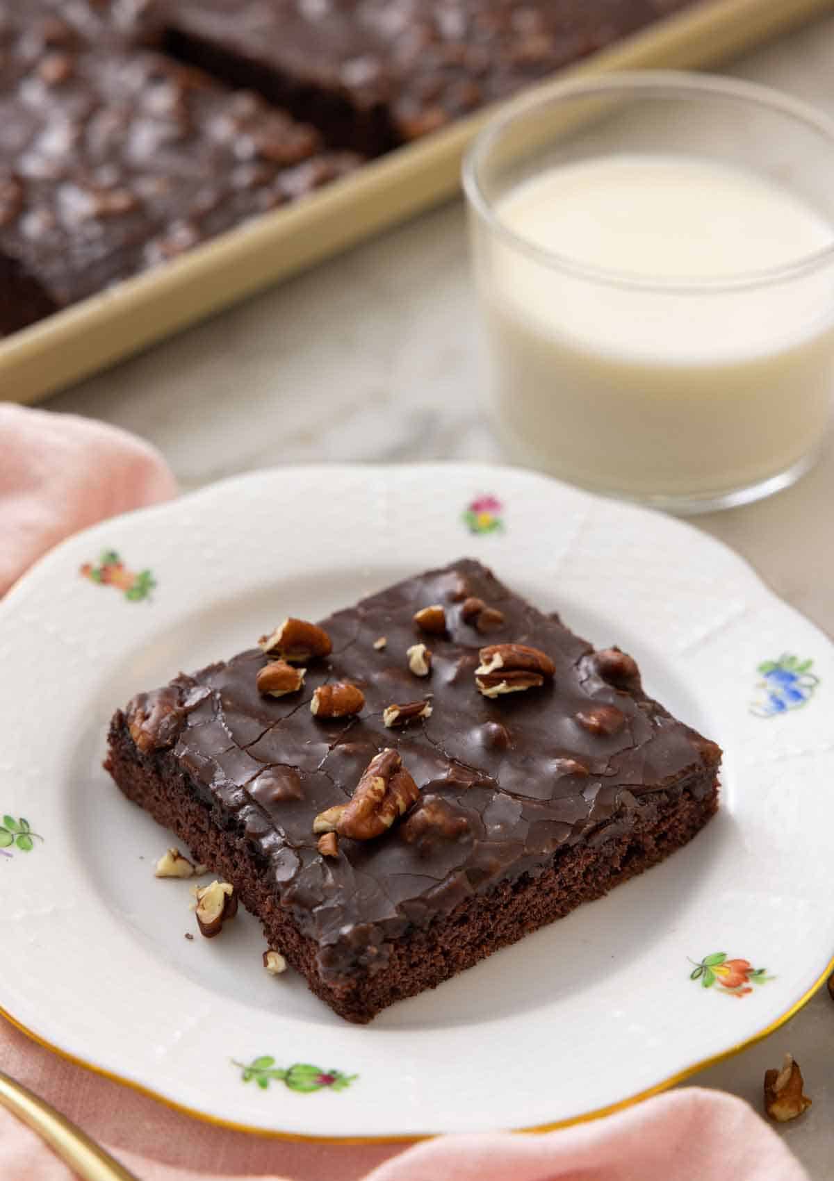 A plate with a square slice of Texas sheet cake with a glass on milk in the back.