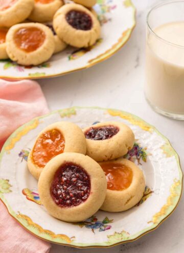A plate with four thumbprint cookies with a glass of milk and another plate of cookies in the back.