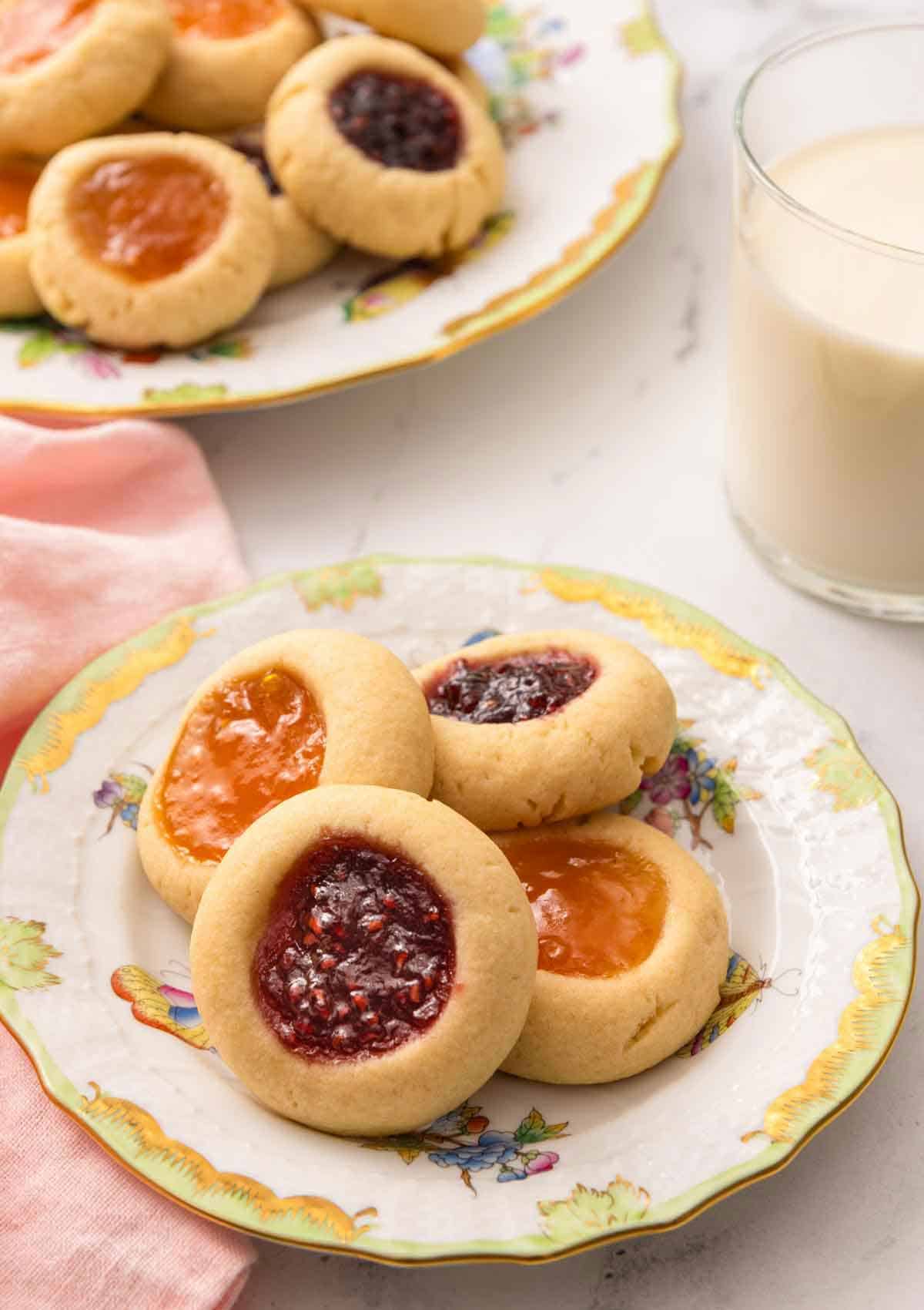 A plate with four thumbprint cookies with a glass of milk and another plate of cookies in the back.