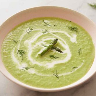 A bowl of asparagus soup with cream drizzled on top with some fresh dill and asparagus garnished on top.