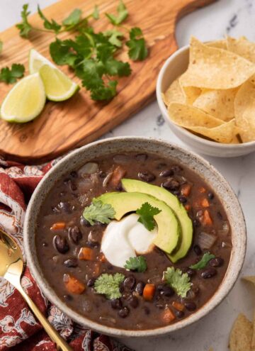 A bowl of black bean soup with sour cream and sliced avocado as garnish. A bowl of chips, lime wedges, and cilantro in the back.