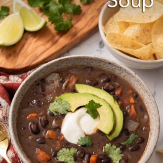 Pinterest graphic of a bowl of black bean soup garnished with cilantro, sour cream, and avocado with a bowl of chips in the background.