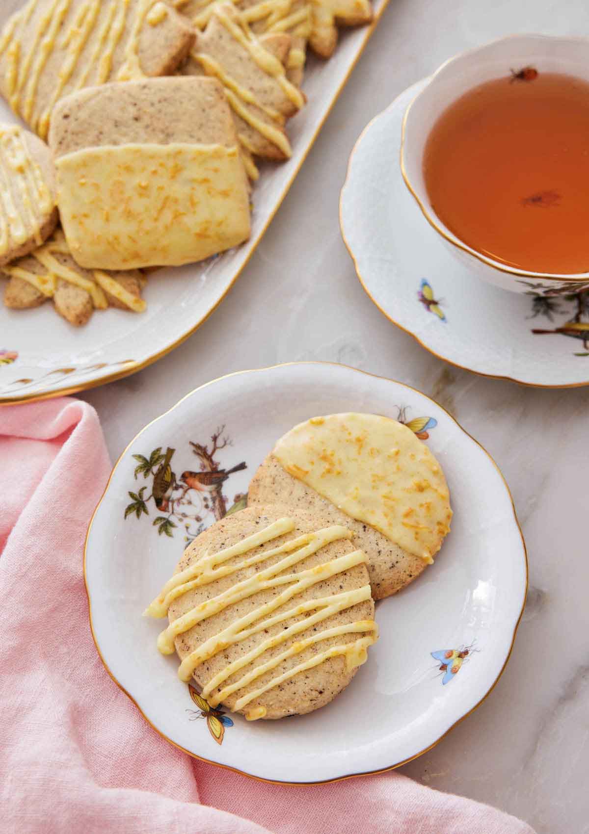 A plate with two browned butter-earl grey cookies by a cup of tea and more cookies in the back.