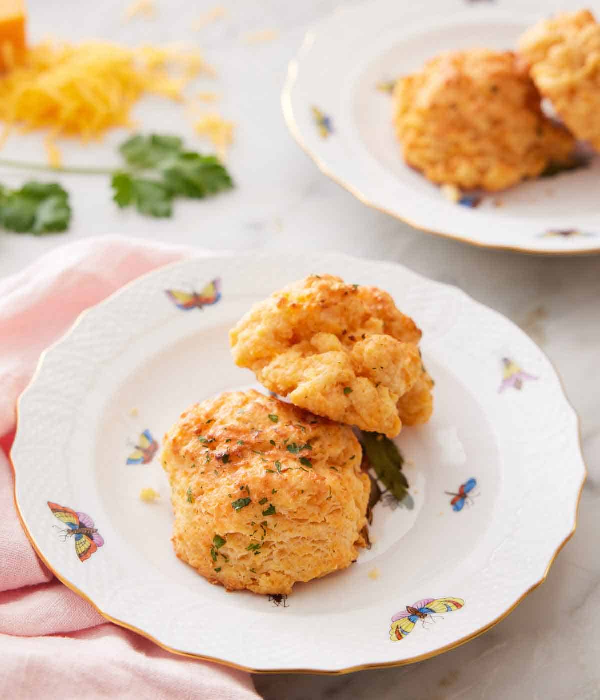 A plate with two cheddar biscuits with more in the back and some shredded cheese and parsley.
