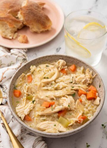A bowl of chicken and rice soup with iced lemon water and bread in the background.