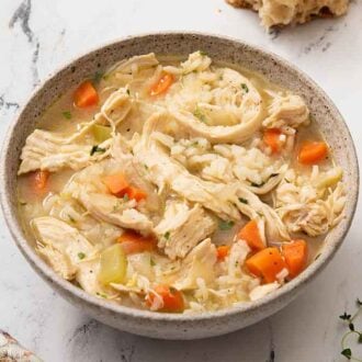 A bowl of chicken and rice soup with bread in the background and fresh thyme on the counter.