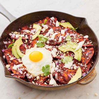 A cast iron skillet with chilaquiles topped with sliced avocado, cilantro, and a fried egg.
