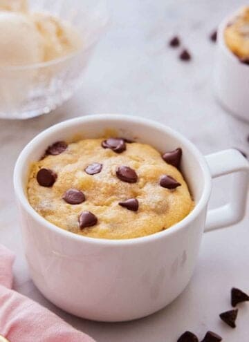 A cookie in a mug with chocolate chips scattered around.