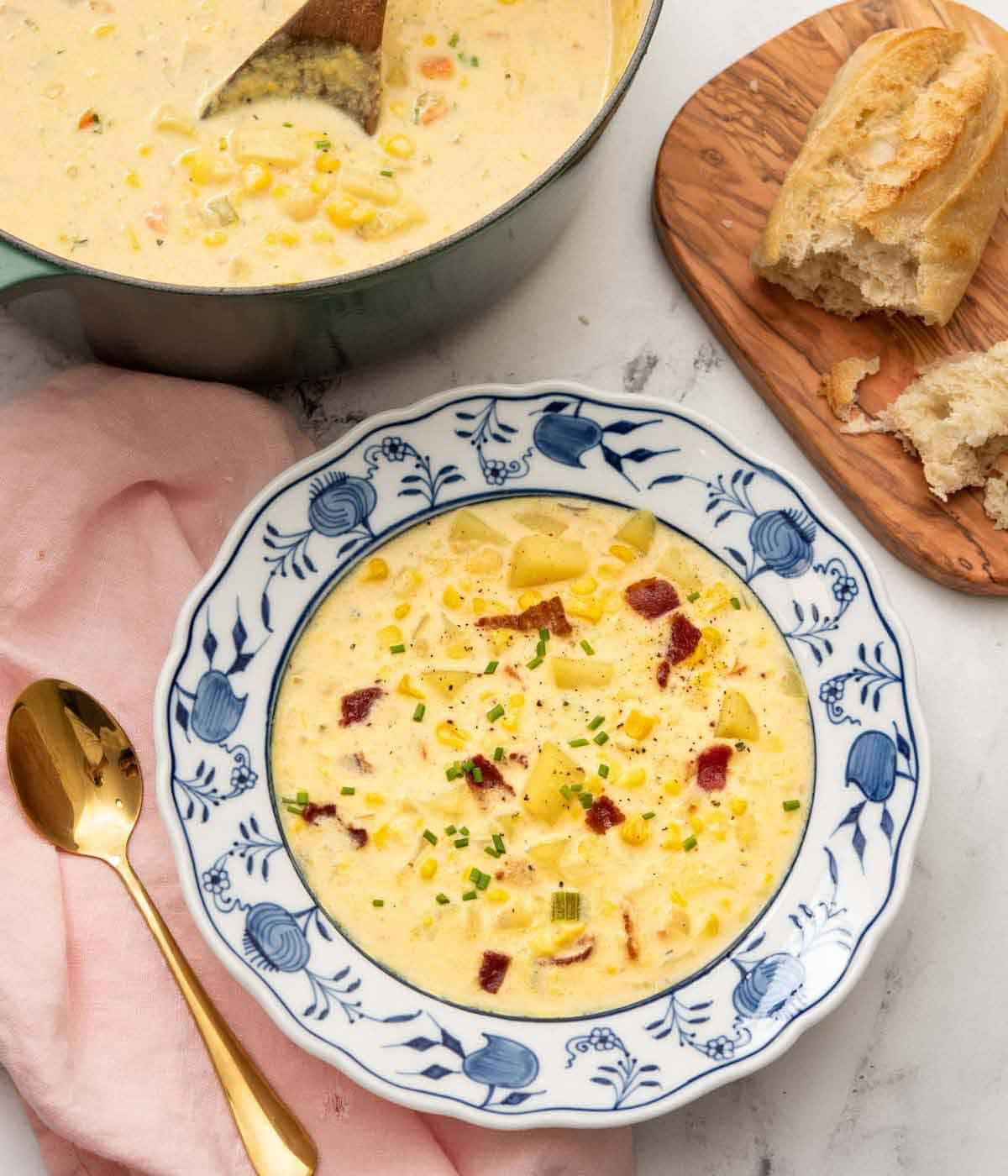 Overhead view of a bowl of corn chowder beside a pot and some crust bread on a serving bowl.