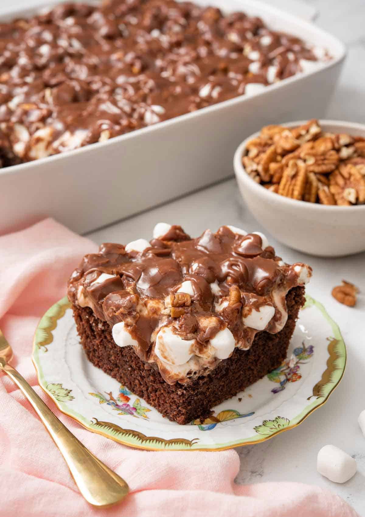 A plate with a square slice of Mississippi mud cake with the rest of the cake in the background.