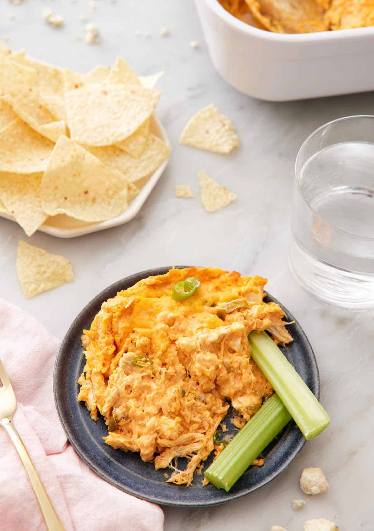 A plate with buffalo chicken dip and two celery sticks with a plate of tortilla chips and a cup of water in the background.