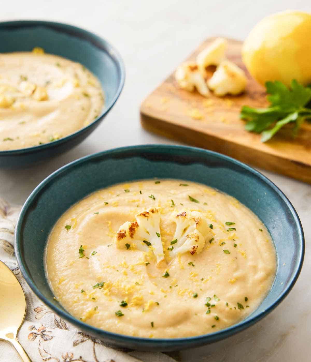 A bowl of cauliflower soup garnished with a roasted cauliflower floret, parsley, and lemon zest with a second bowl and garnishes on a cutting board in the background.