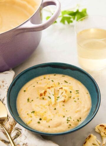 A bowl of cauliflower soup topped with a roasted cauliflower floret in front of a glass of wine and a dutch oven.