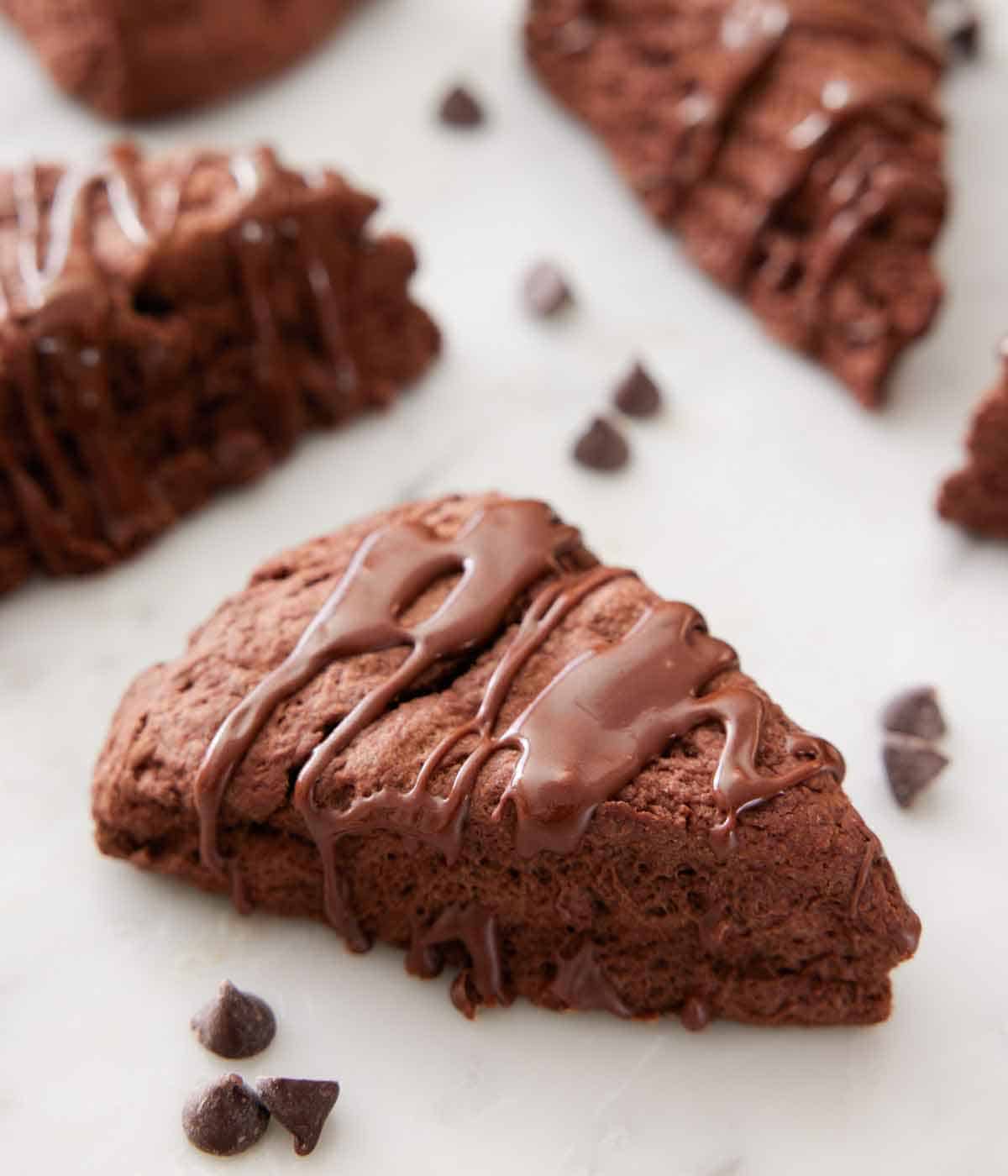 A chocolate scone with chocolate drizzled over on top with some more scones and chocolate chips scattered around.