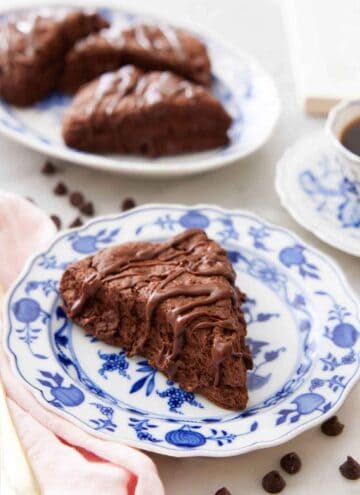 A plate with a chocolate scone with chocolate drizzled on top with a platter of scones in the background.